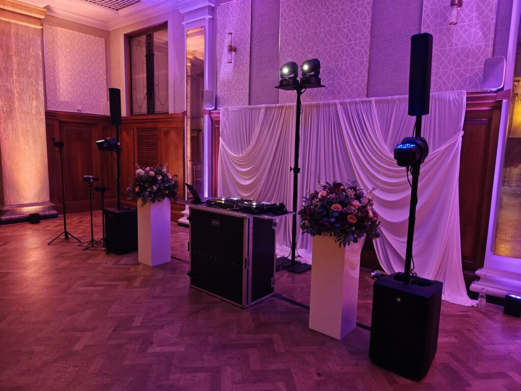 DJ booth, column speakers and moving head lights set up in front of white drapes in a ballroom