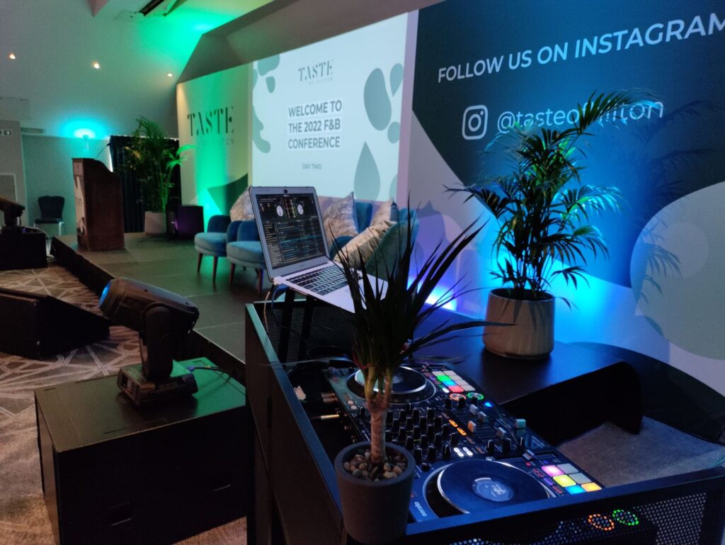 DJ controller and laptop beside a conference stage, with a moving head light and green and blue uplighting