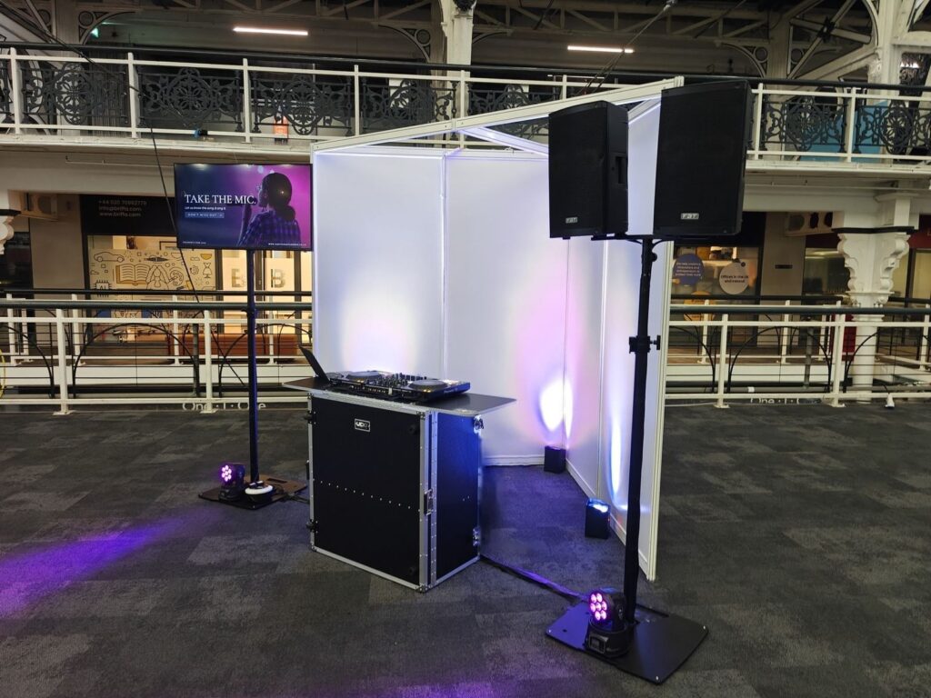 DJ booth, two speakers on a stand, a TV on a stand and moving head lights set up on an exhibition hall floor