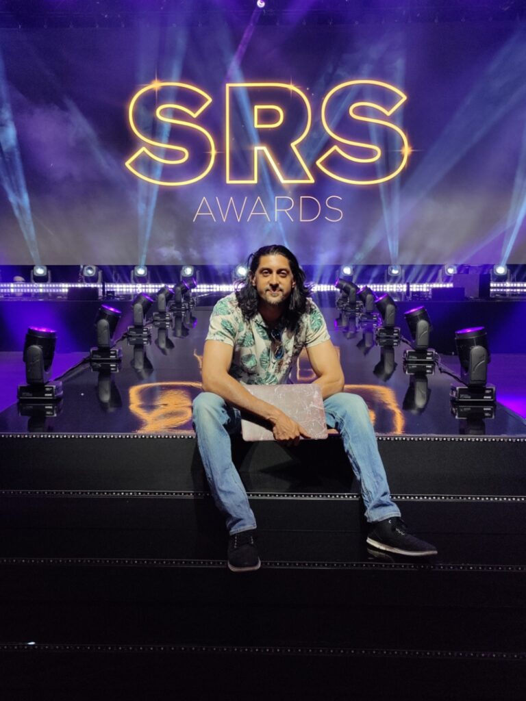 DJ SR sitting on the stage steps in front of the SRS Awards screen, 2023